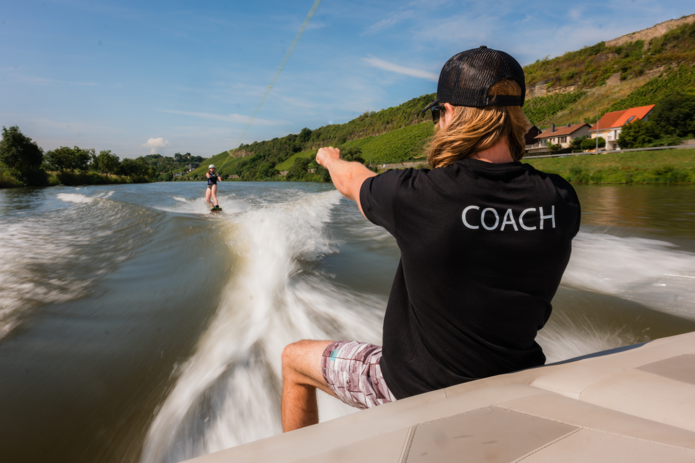 Image d'un animateur de wakeboarding assis sur la partie arrière d'un bâteau en train de donner des instructions à un enfant qui est en train de faire du wakeboard derrière la bâteau