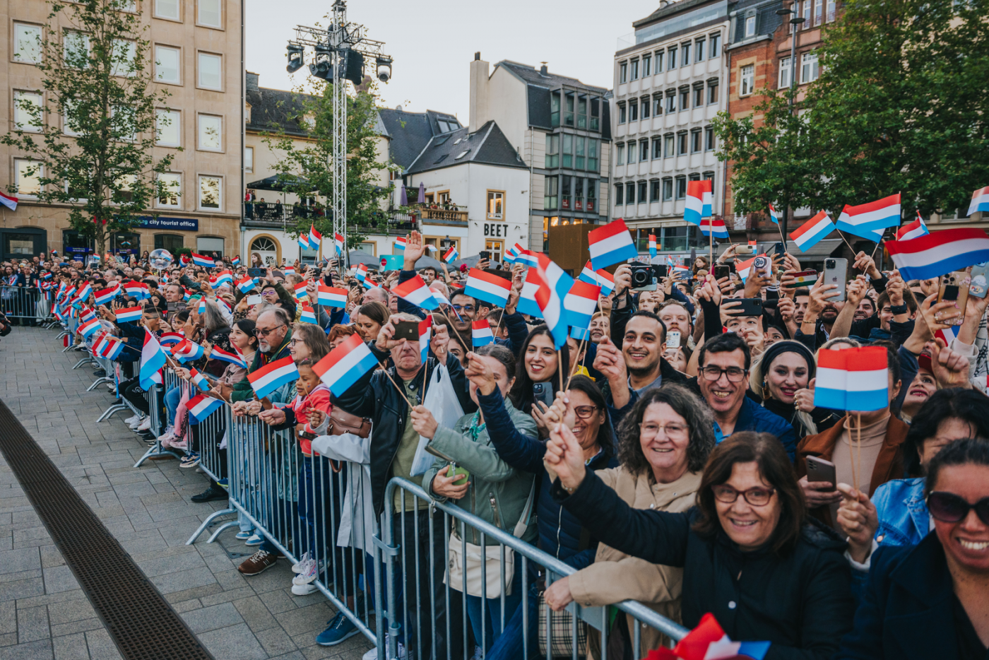 Une grande foule joyeuse rassemblée derrière des barrières métalliques agite des drapeaux luxembourgeois et sourit en direction de l’objectif lors d’un événement public sur une place urbaine. Beaucoup tiennent leur téléphone pour capturer le moment. Les bâtiments environnants et l’ambiance festive évoquent une célébration nationale ou civique à Luxembourg-ville.